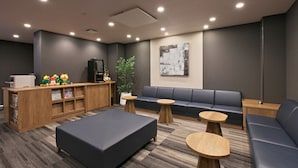 A modern lounge area with a sofa, ottoman, and small tables, a reception desk with a plant, and a large abstract painting on the wall. - Hotel Route Inn Toyama Ekimae (Toyama)