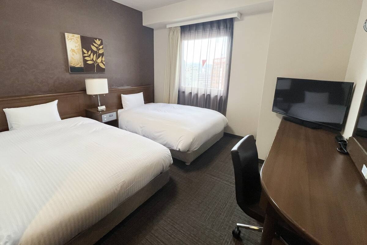 A hotel room with two beds, a desk with a TV, a lamp, and a window with curtains.