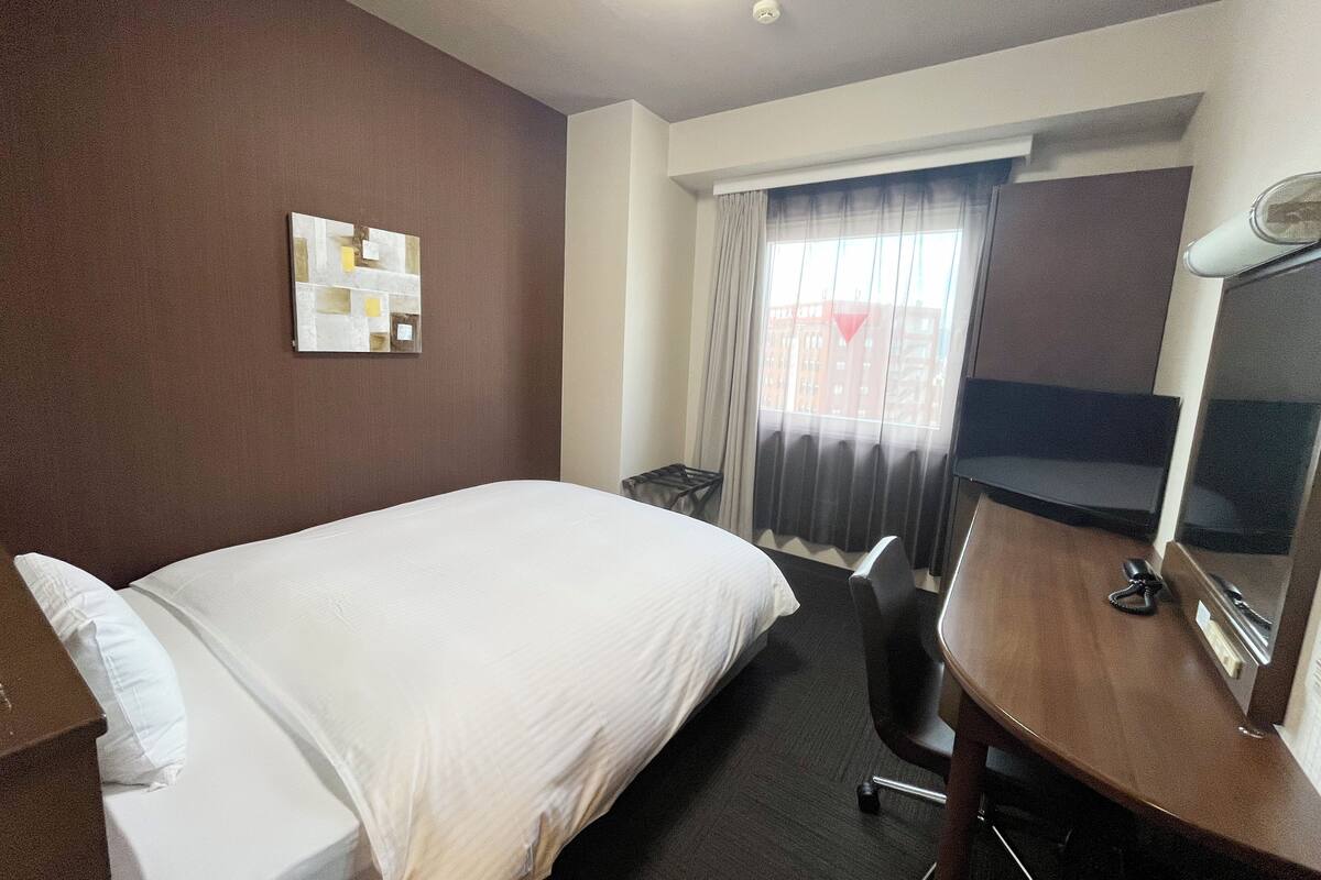A hotel room with a bed, a desk, a chair, a flat-screen TV, a ceiling light, and a window with curtains.