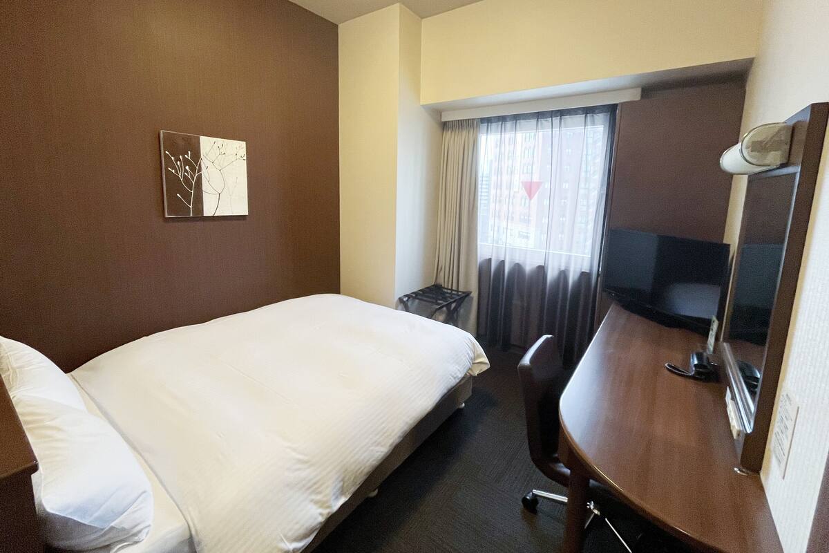 A hotel room with a bed, a desk, a chair, a small table, a mirror, and a window with curtains.