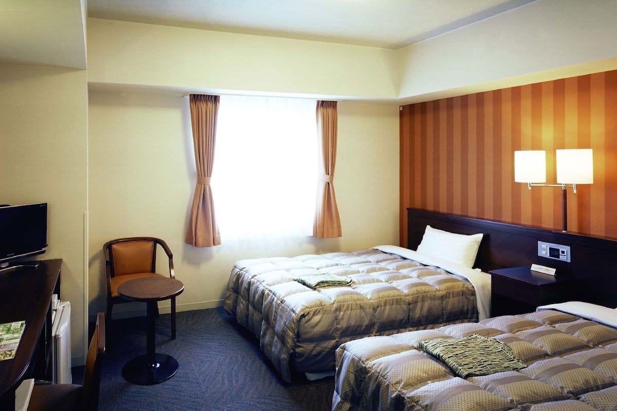 Habitación de hotel con dos camas, un escritorio, una silla, un televisor y una ventana con cortinas.