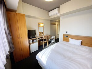 A hotel room with a bed, a desk with a computer, a television, a wardrobe, and an air conditioning unit.