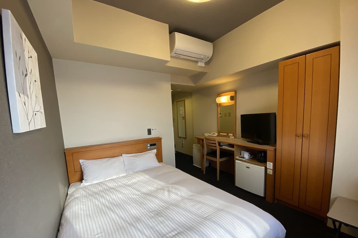 Ett hotellrum med en säng, ett skrivbord, en stol, en tv, en garderob och en luftkonditioneringsanläggning.