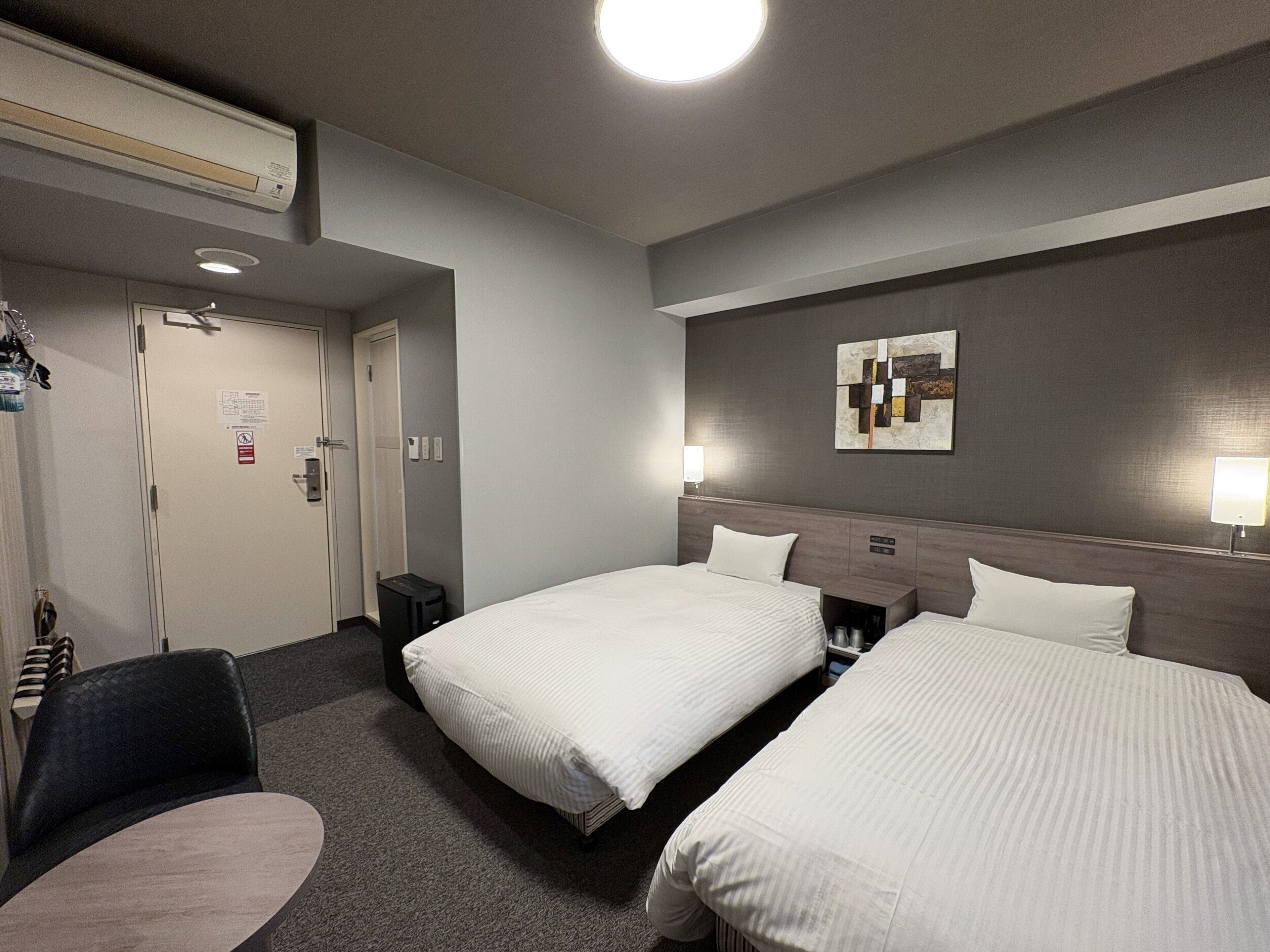 Ein Hotelzimmer mit zwei Betten, einem Schreibtisch, einem Stuhl, einem kleinen Tisch, einem Wandbild und einer Deckenleuchte.