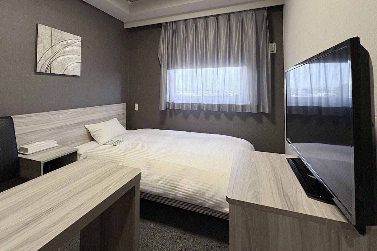 A hotel room with a bed, a desk, a chair, a TV, and a window with curtains.