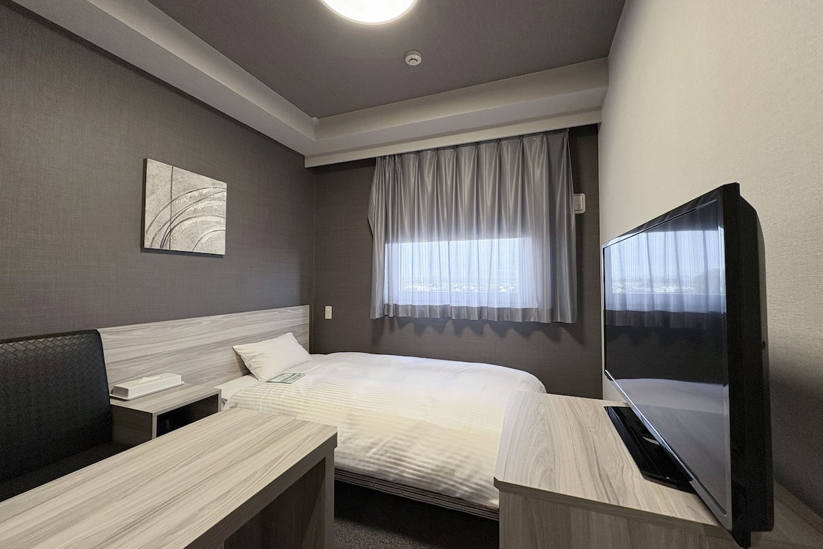 A modern hotel room with a bed, a desk, a chair, a flat-screen TV, a window with curtains, and a ceiling light.