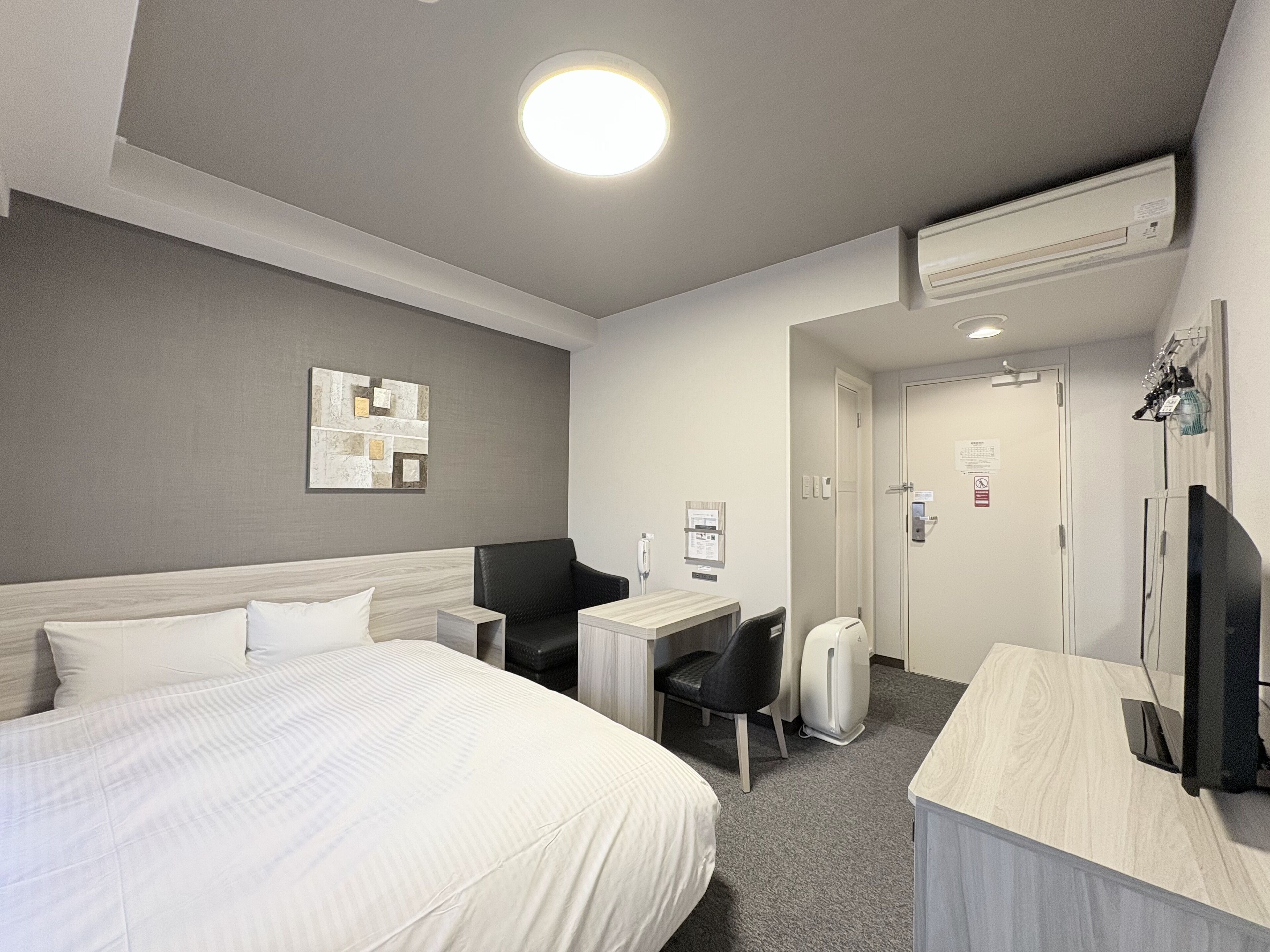 A hotel room with a bed, a desk, a chair, a TV, and an air conditioning unit.