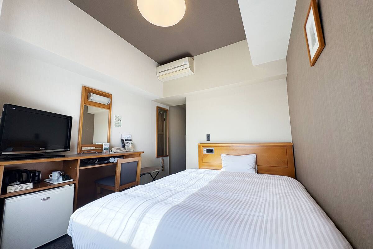A hotel room with a bed, a desk with a computer, a TV, a mirror, a coffee machine, and an air conditioner.
