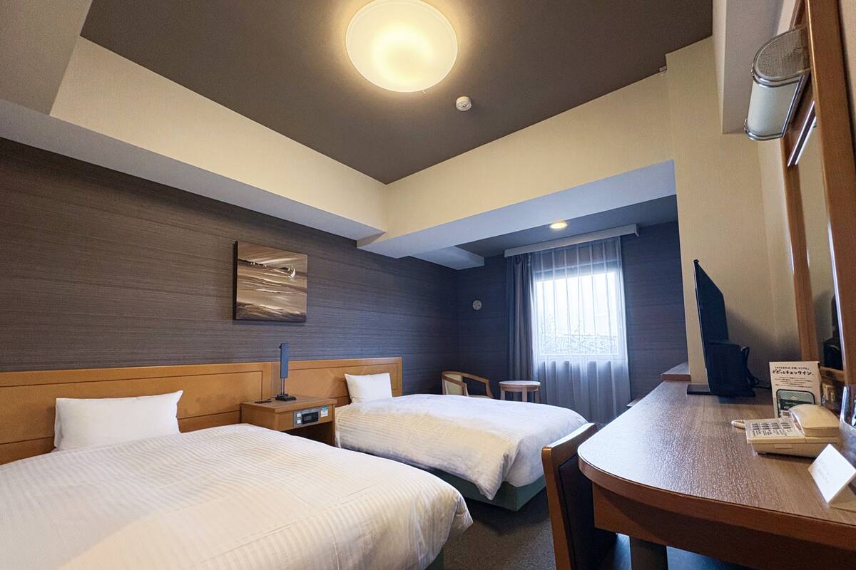 A hotel room with two beds, a desk, a chair, a clock, and a window with curtains.