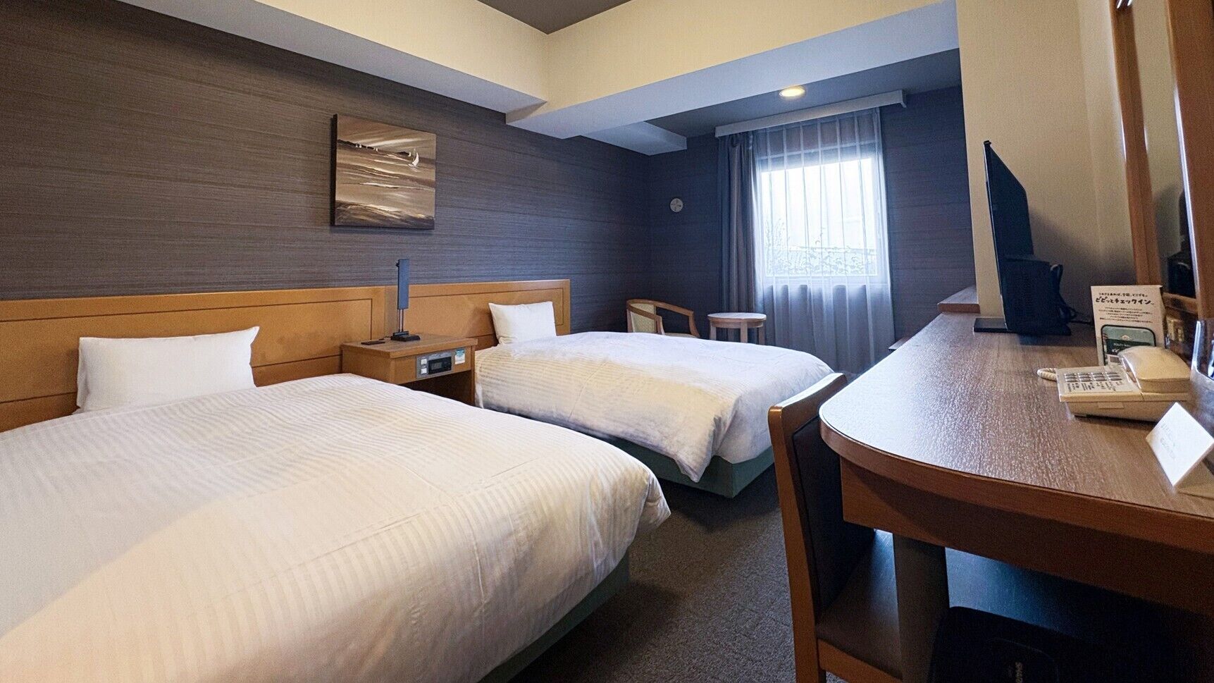 A hotel room with two beds, a desk, a chair, a clock, and a window with curtains.