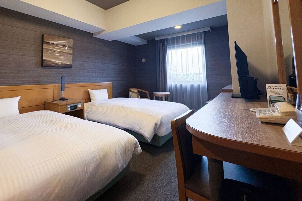 A hotel room with two beds, a desk, a chair, a clock, and a window with curtains.