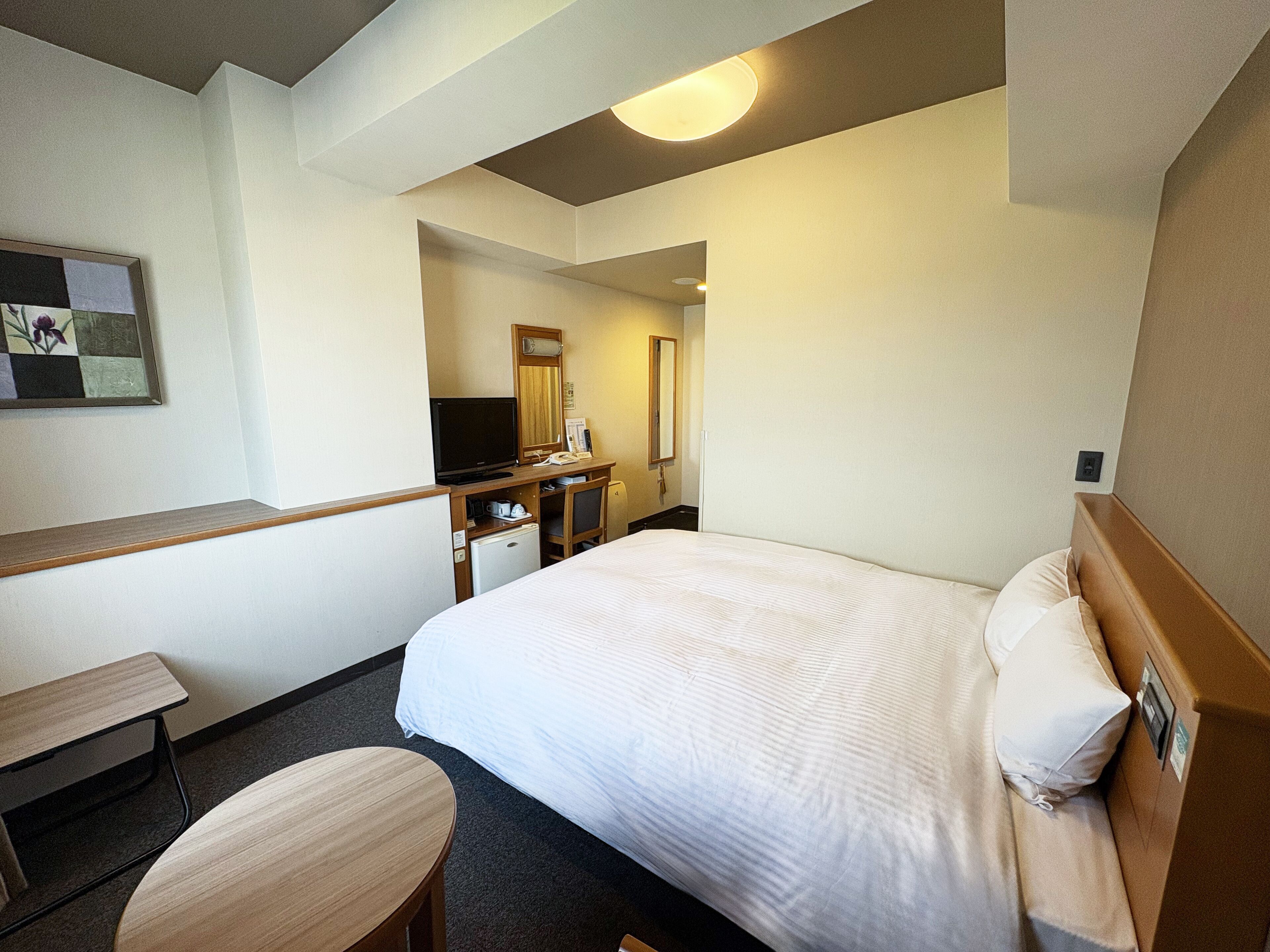 A hotel room with a bed, a desk, a chair, a television, a refrigerator, and a mirror.