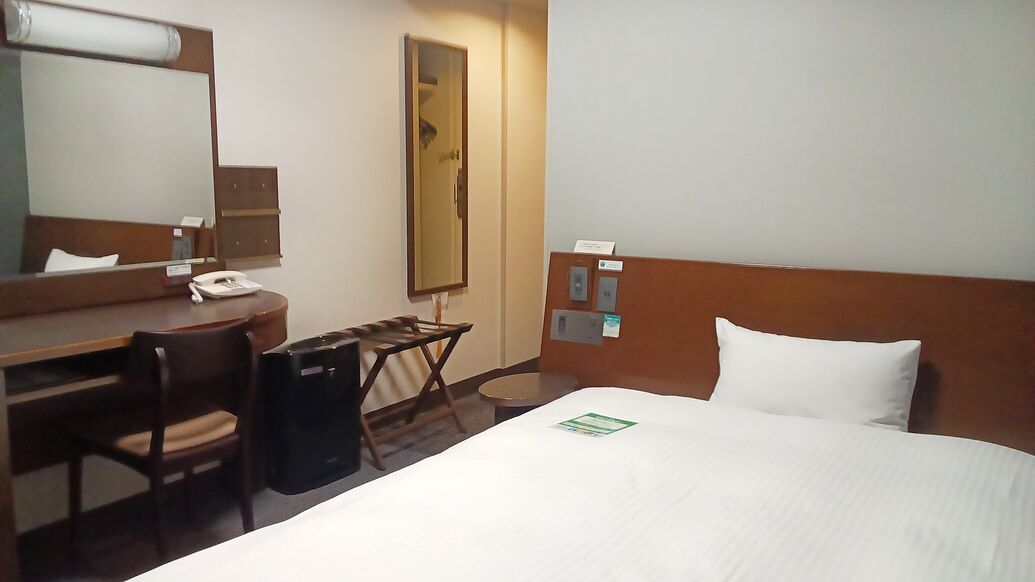 A hotel room with a bed, a desk, a chair, a mirror, and a trash bin.