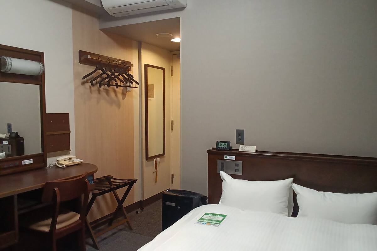 Une chambre d’hôtel comprenant un lit, un bureau avec un téléphone, une chaise, un miroir, un porte-manteau et une horloge.
