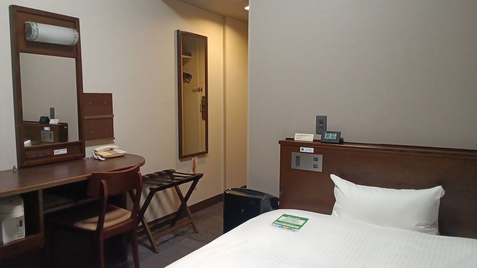 A hotel room with a bed, a desk with a chair, a mirror, a clock, and a small trash bin.