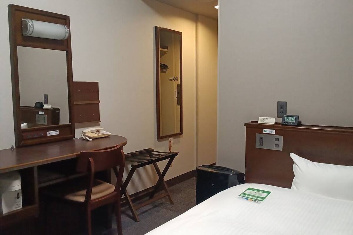 Une chambre d’hôtel comprenant un lit, un bureau avec une chaise, un miroir, une horloge et une petite poubelle.