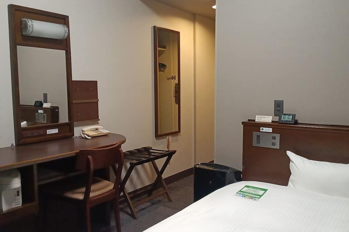 Una habitación de hotel con una cama, un escritorio con una silla, un espejo, un reloj y un pequeño cubo de basura.
