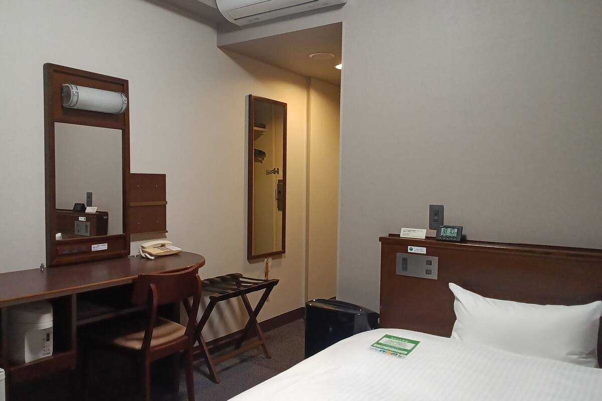 Une chambre d’hôtel comprenant un lit, un bureau avec une chaise, un miroir, une horloge et une poubelle.