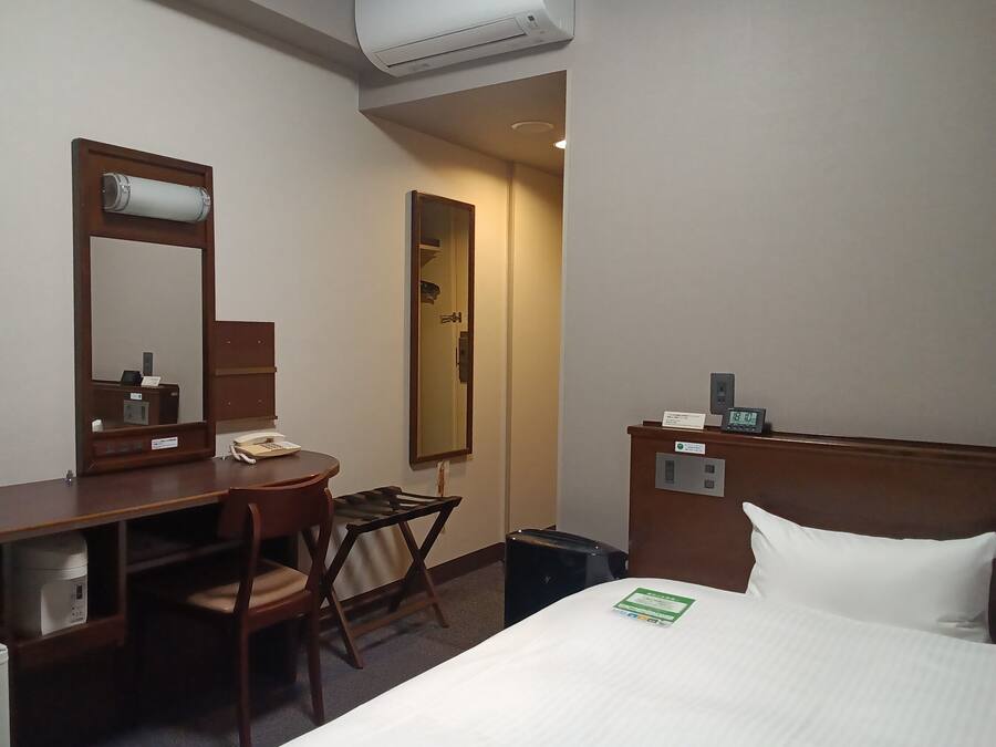 A hotel room with a bed, a desk with a chair, a mirror, a clock, and a trash bin.