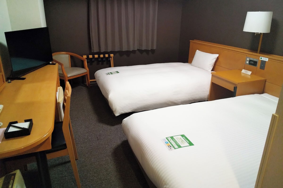 A hotel room with two single beds, a desk with a chair, a TV, a lamp, and a window with curtains.