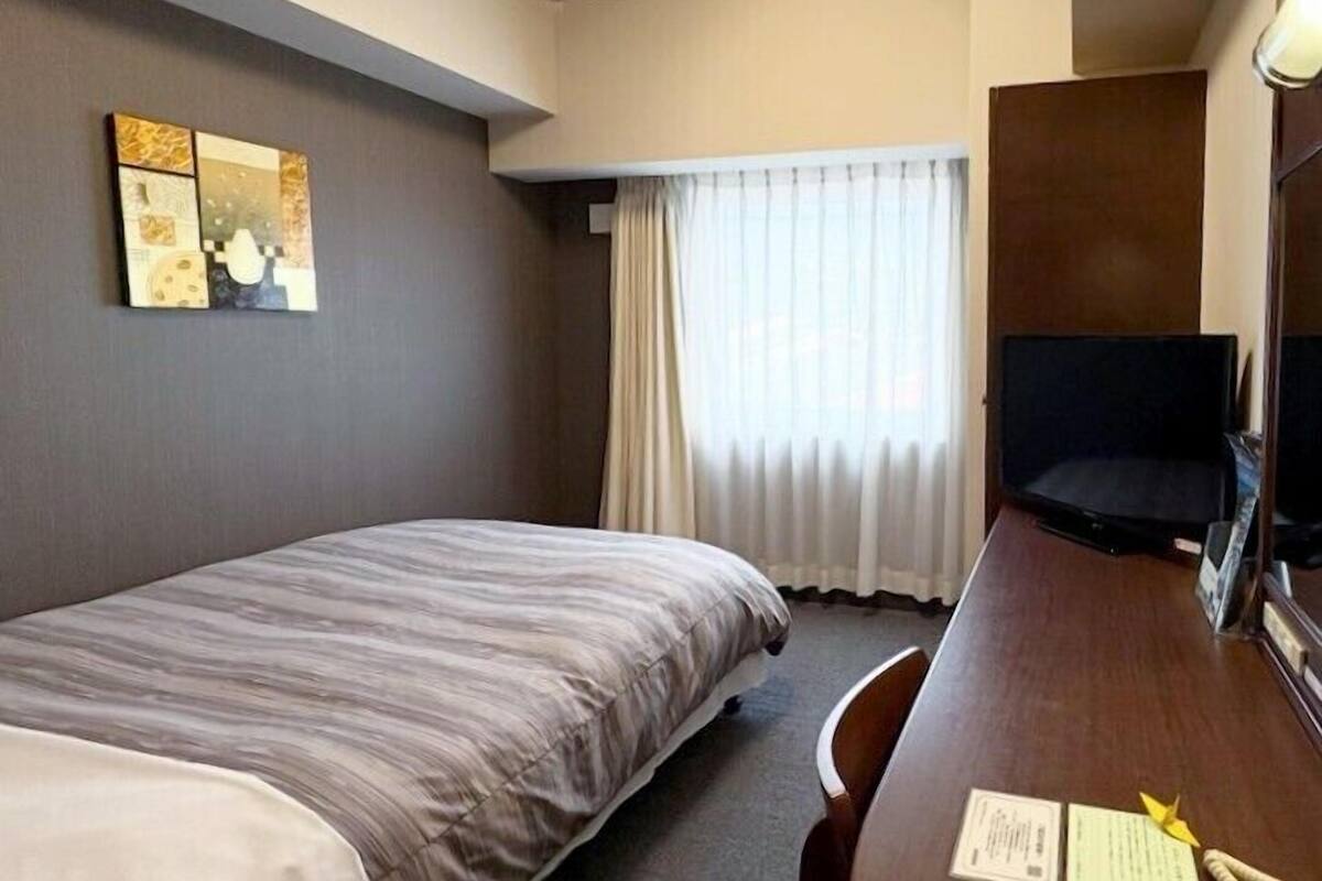 Ein Hotelzimmer mit Bett, Schreibtisch mit Stuhl, Fernseher und einem Fenster mit Vorhängen.