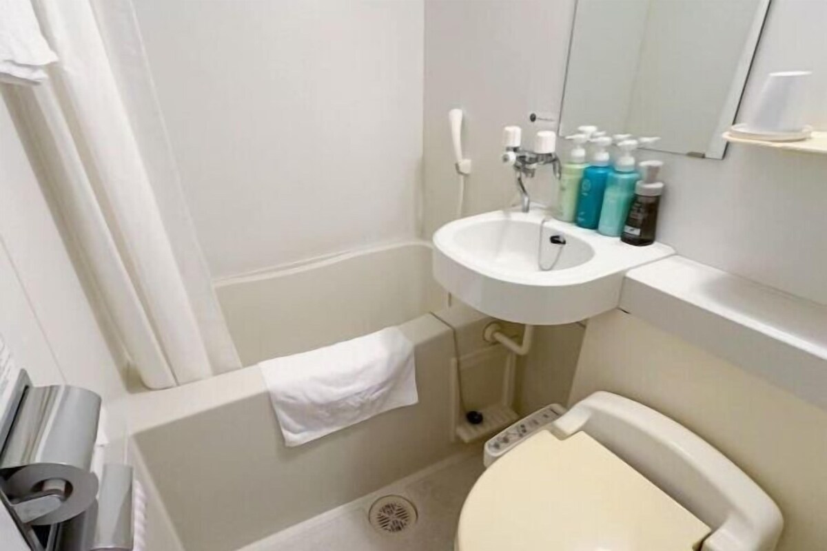 A small, white bathroom with a sink, toilet, and a mirror. There are toiletries on the sink and a towel hanging on the wall.