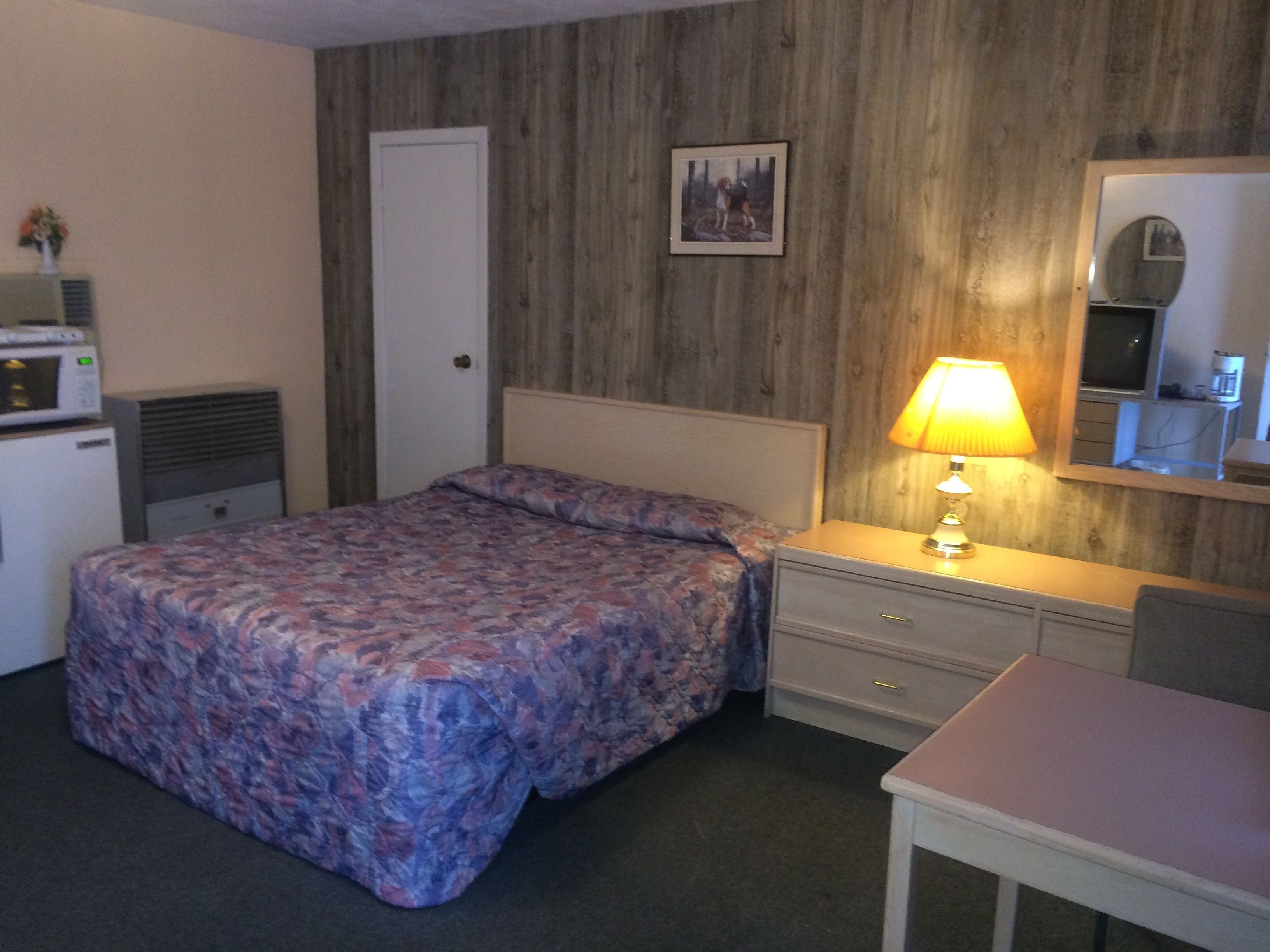 Une chambre d’hôtel comprenant un lit, une table de chevet avec une lampe, un petit bureau, un miroir, une porte et une kitchenette équipée d’un four à micro-ondes.