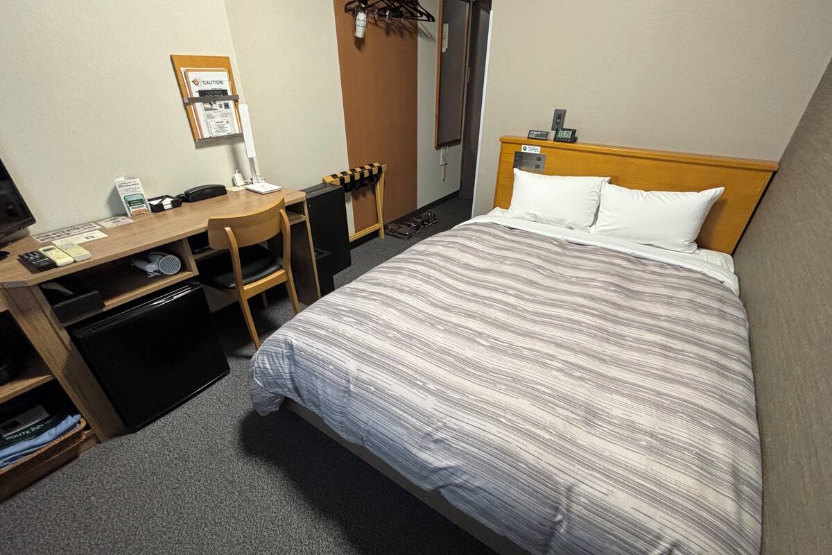 A hotel room with a bed, a desk, a chair, a small table, and a coat rack.