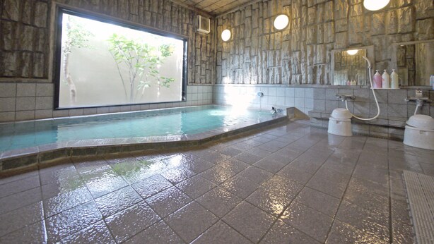 A spa area with a large soaking tub, a shower area, and a stone wall with a window overlooking greenery. - Hotel Route-Inn Court Fujioka (Fujioka)