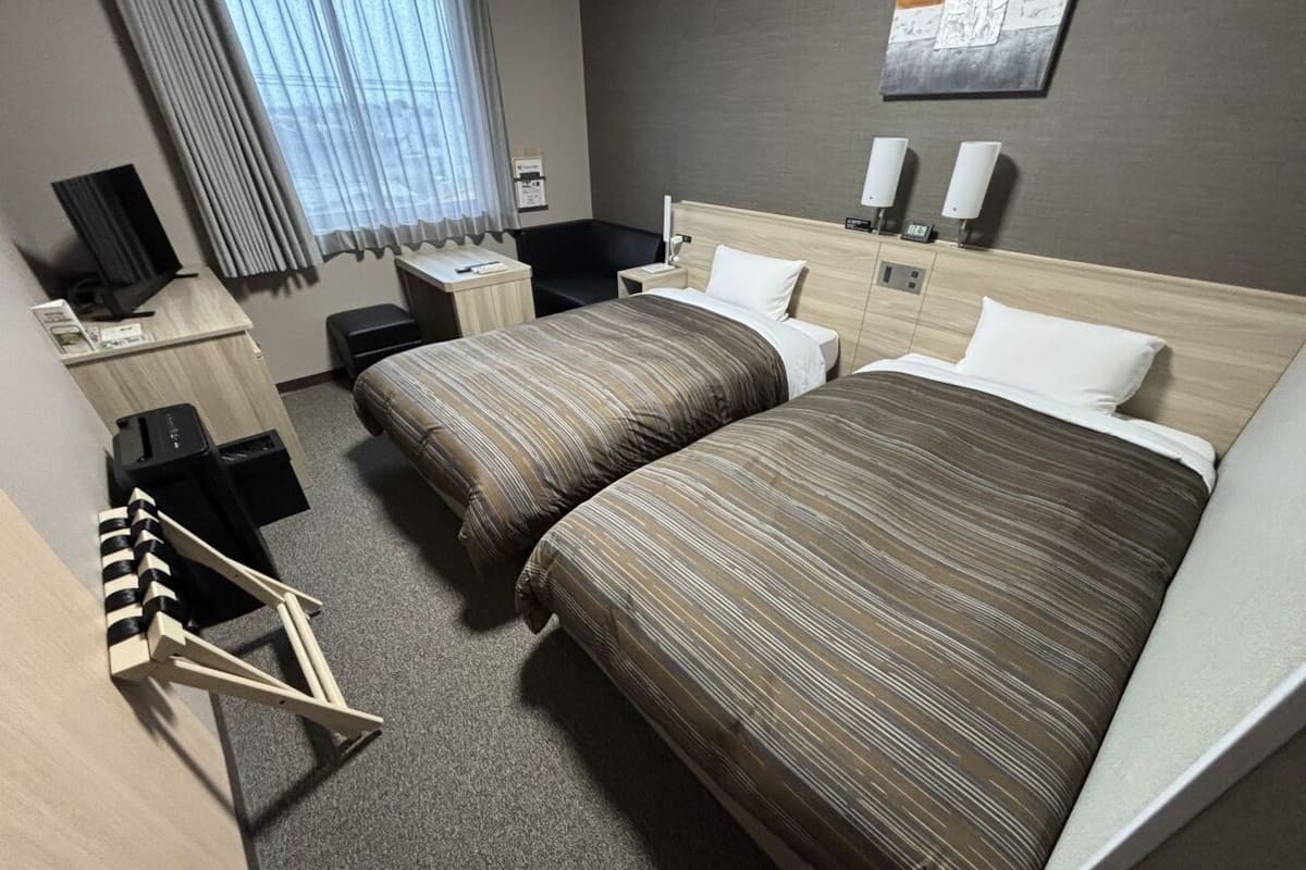 A hotel room with two beds, a desk, a chair, a TV, and a window with curtains.
