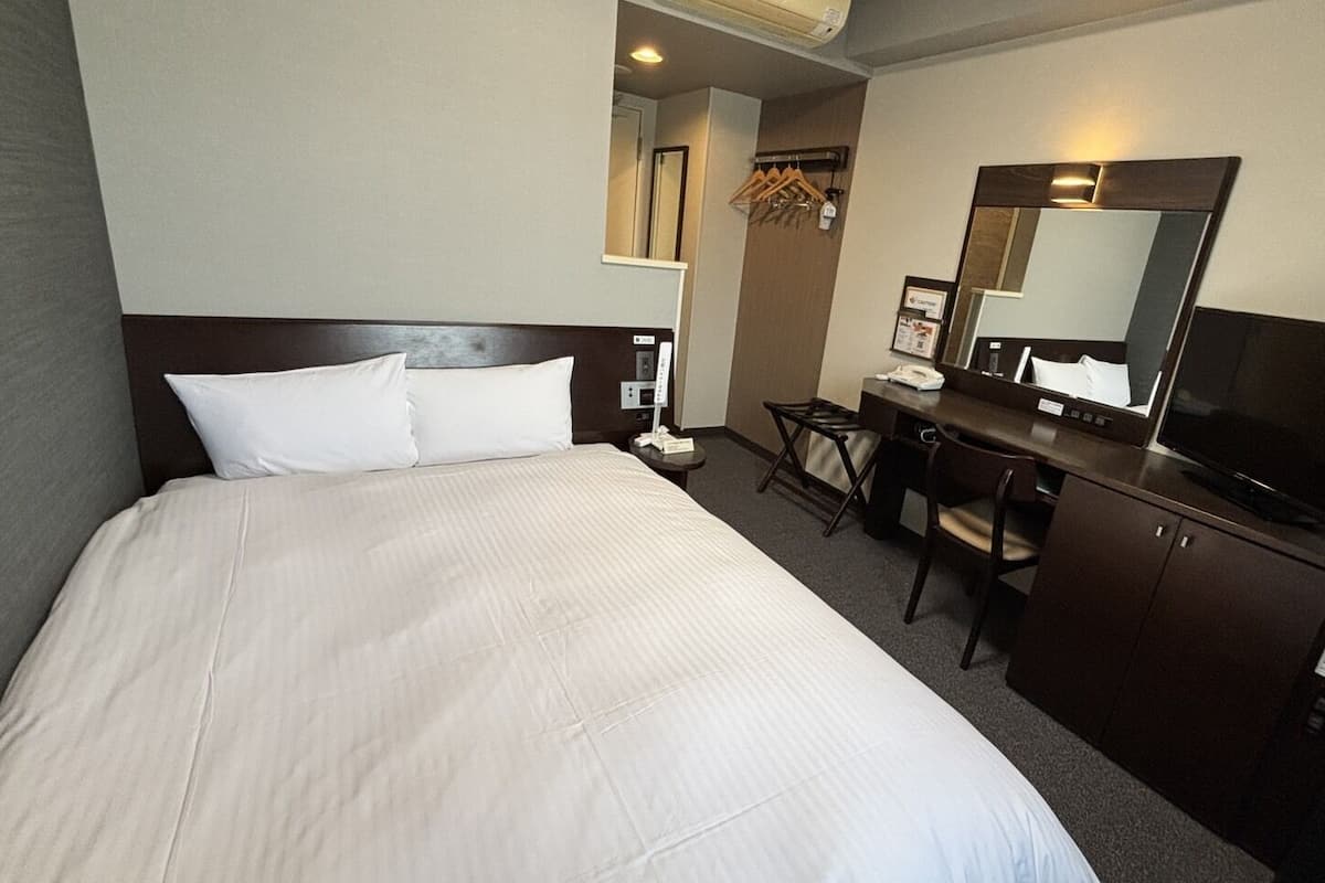 A hotel room with a large bed, a desk with a chair, a television, a mirror, and a wardrobe.