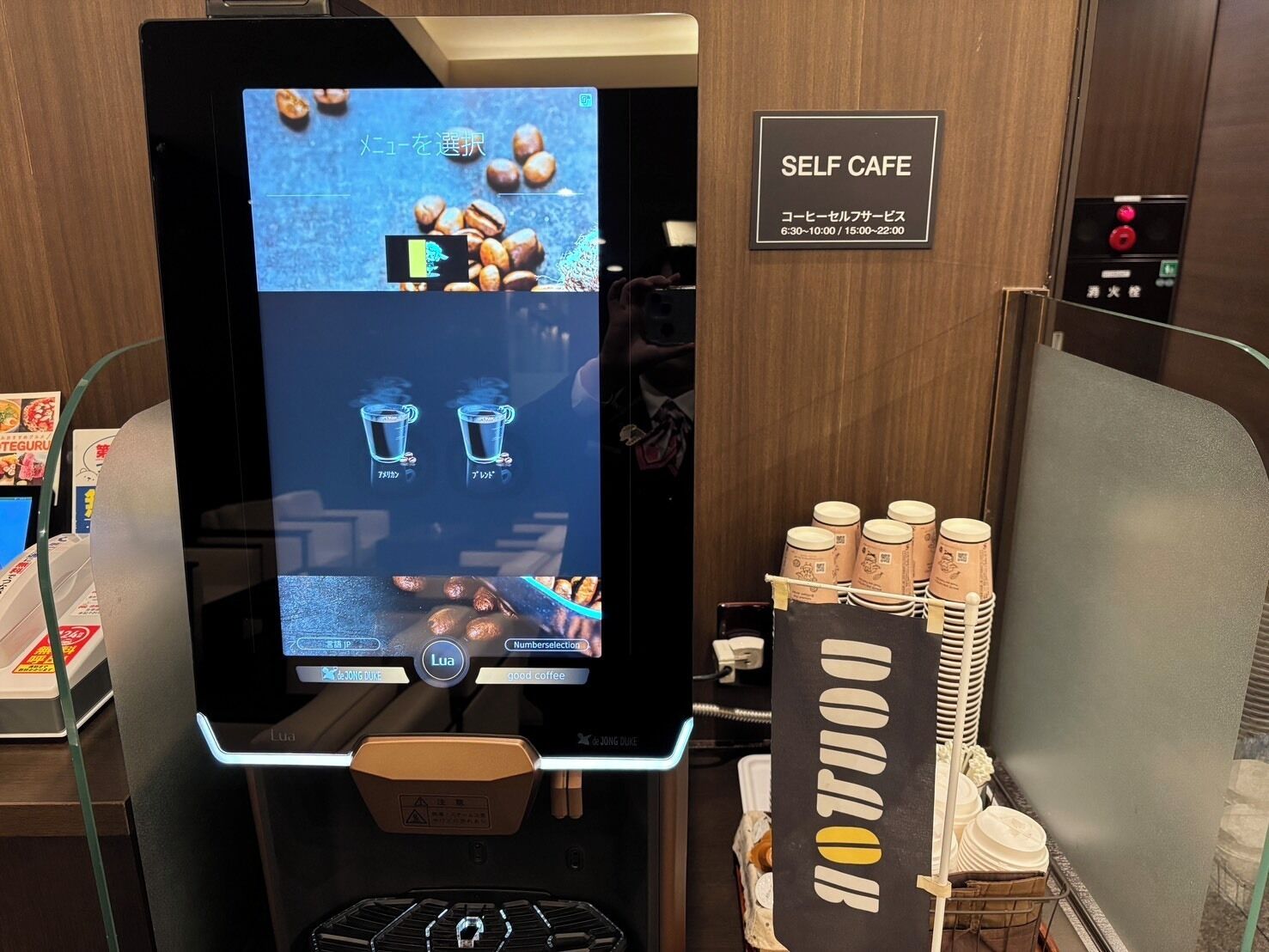 Uma máquina de café automática com interface digital, um aviso com o horário de funcionamento e uma pilha de copos descartáveis.