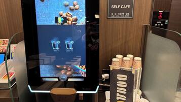 Uma máquina de café automática com interface digital, um aviso com o horário de funcionamento e uma pilha de copos descartáveis.