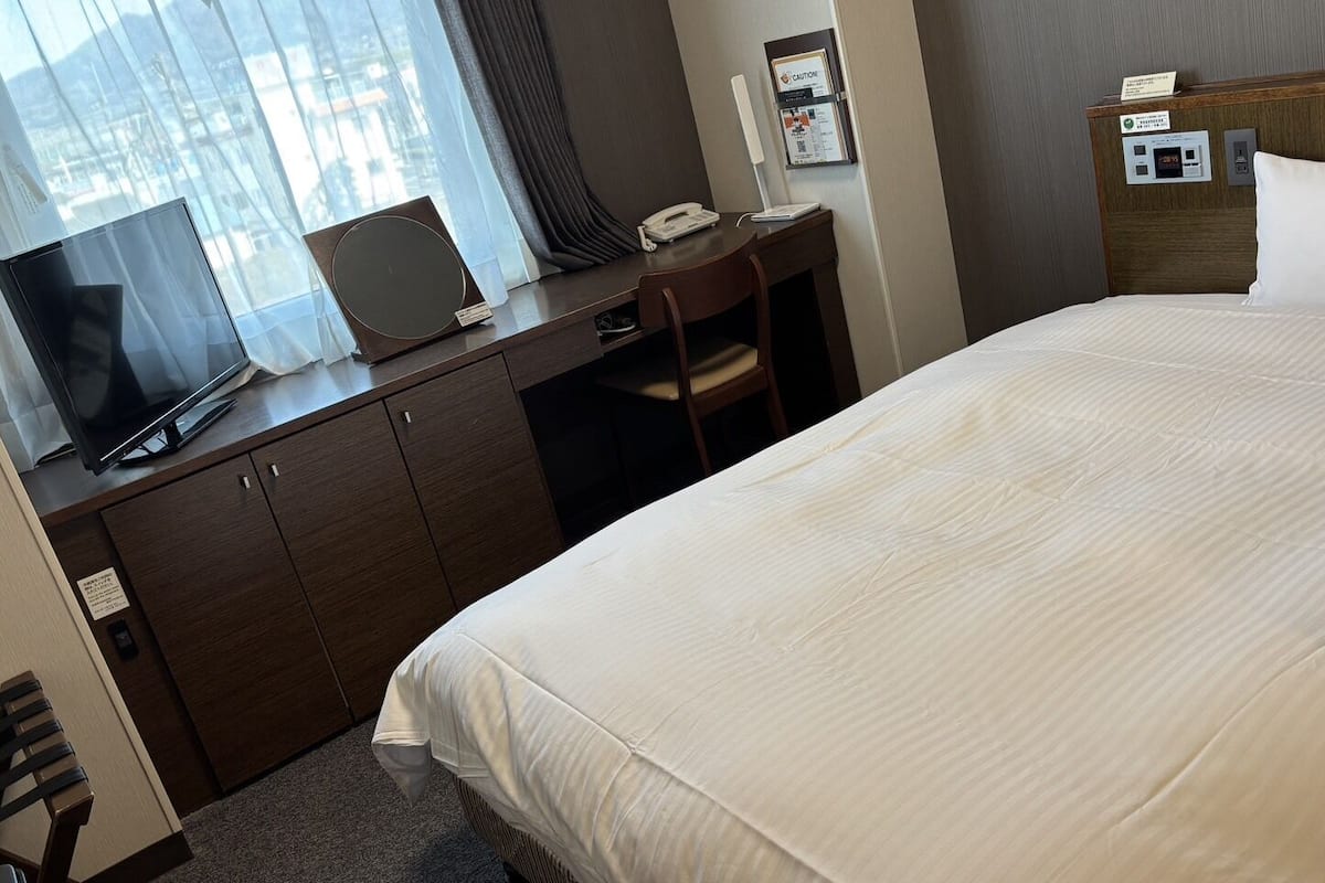 A hotel room with a bed, a desk with a laptop, a telephone, and a nightstand with a lamp.