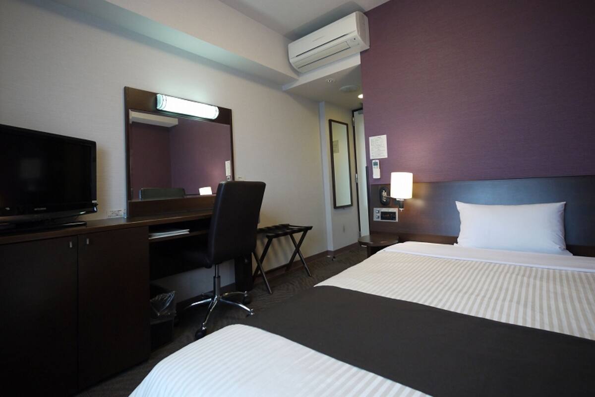 Habitación de hotel con cama, escritorio, silla, televisor, unidad de aire acondicionado y mesita de noche con lámpara.