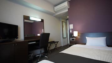 A hotel room with a bed, a desk, a chair, a television, an air conditioning unit, and a nightstand with a lamp.