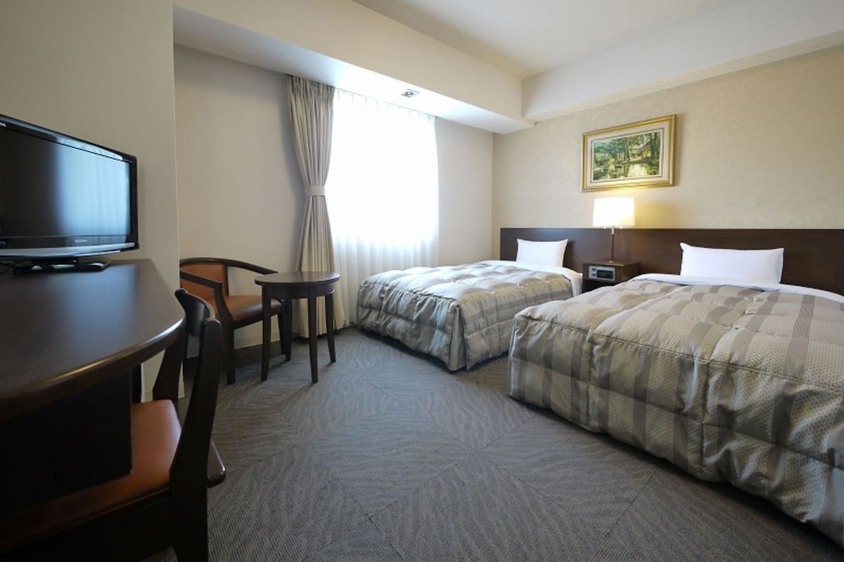Habitación de hotel con dos camas, un escritorio con televisor, una silla, una mesita y un cuadro en la pared.