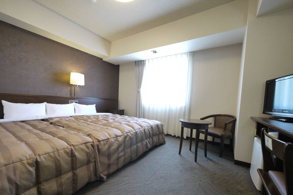 Una habitación de hotel con una cama grande, un escritorio con silla, televisor y ventana con cortinas.