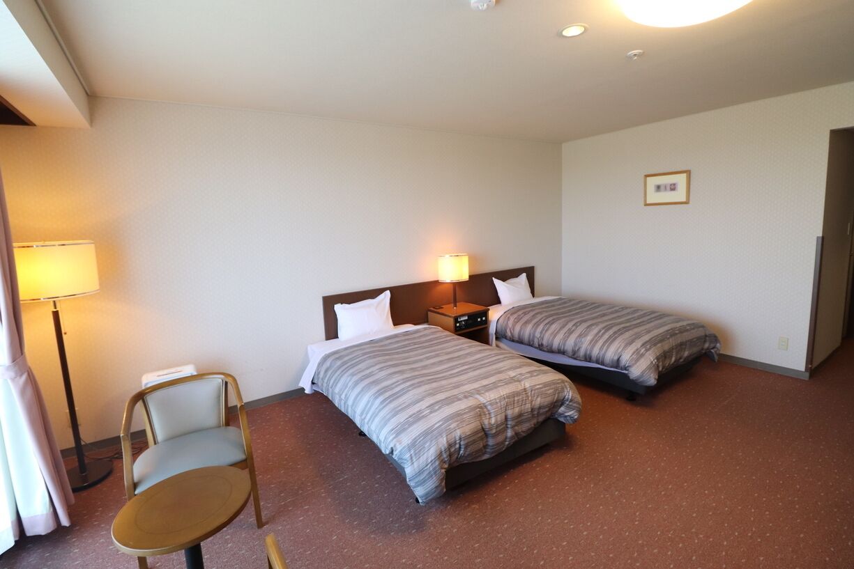 Quarto de hotel com duas camas de solteiro, uma cadeira, uma mesa de cabeceira e um abajur.