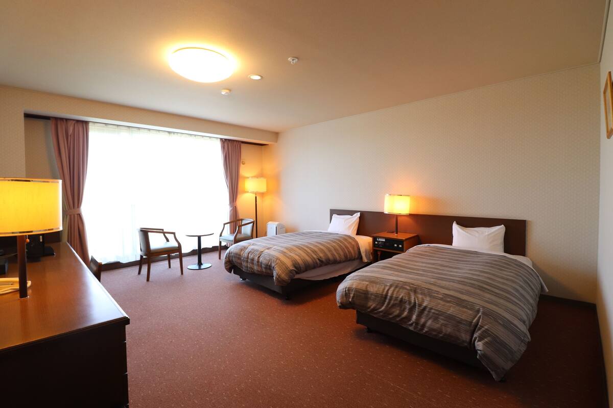 A hotel room with two beds, a desk, a chair, and a lamp.