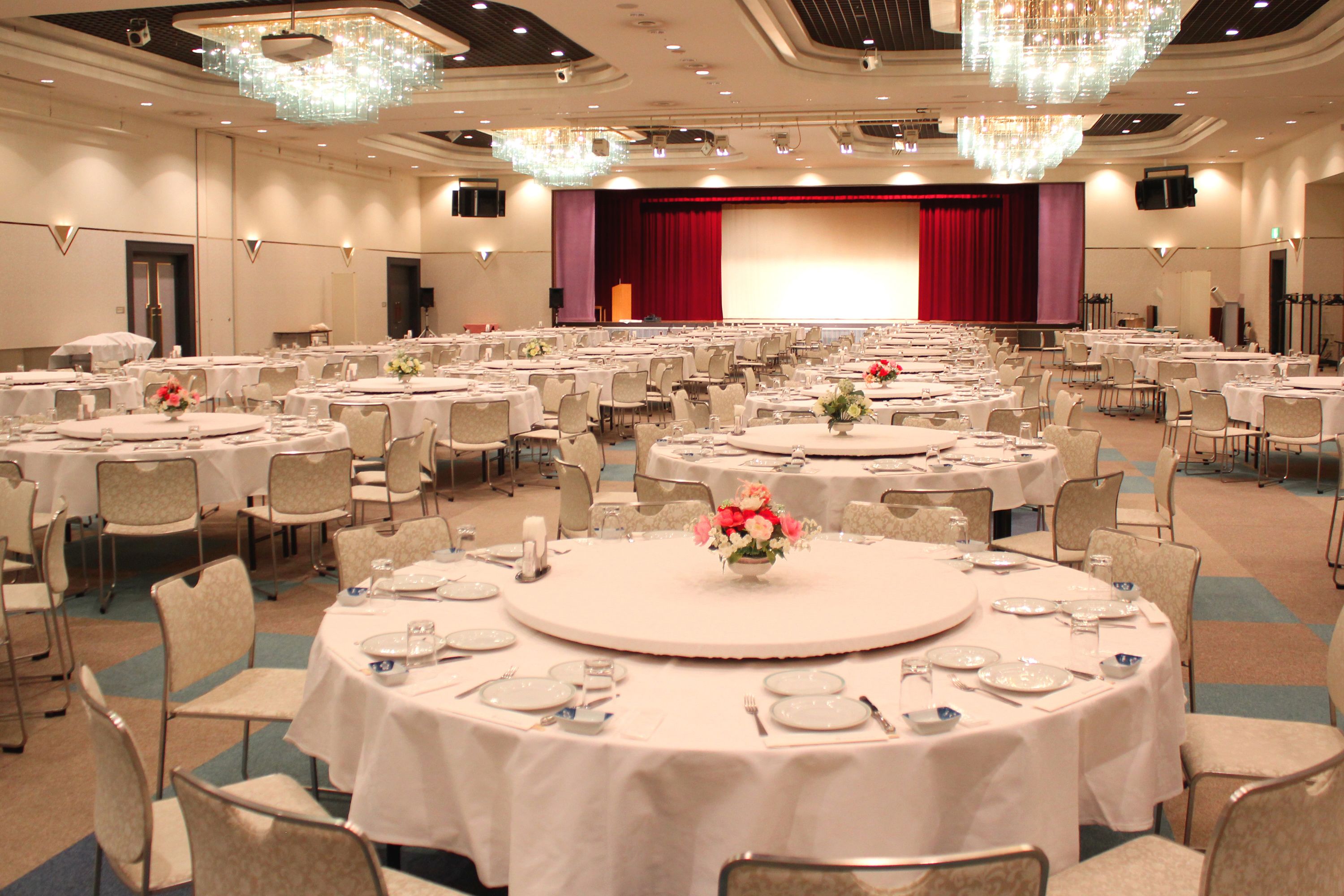 Une grande salle de banquet avec des tables rondes, des chaises et une scène surmontée d’un rideau rouge.