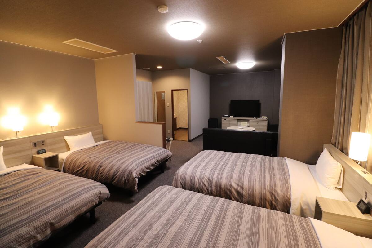 A hotel room with three beds, a desk, a television, and a bathroom visible in the background.