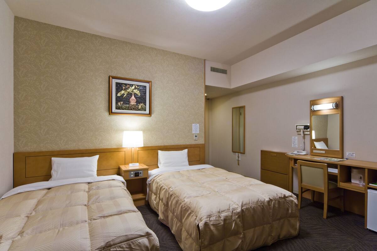 Habitación de hotel con dos camas, un escritorio, una silla, un espejo, un cuadro en la pared y una mesita de noche con una lámpara.