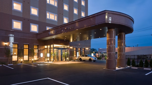 A hotel entrance with a covered walkway, a car parked in front, and a parking lot. - Hotel Route Inn Tomakomai Ekimae (Tomakomai)