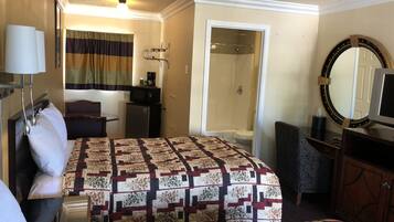 A hotel room with a bed, a desk, a microwave, a refrigerator, a shower, a mirror, and a television.