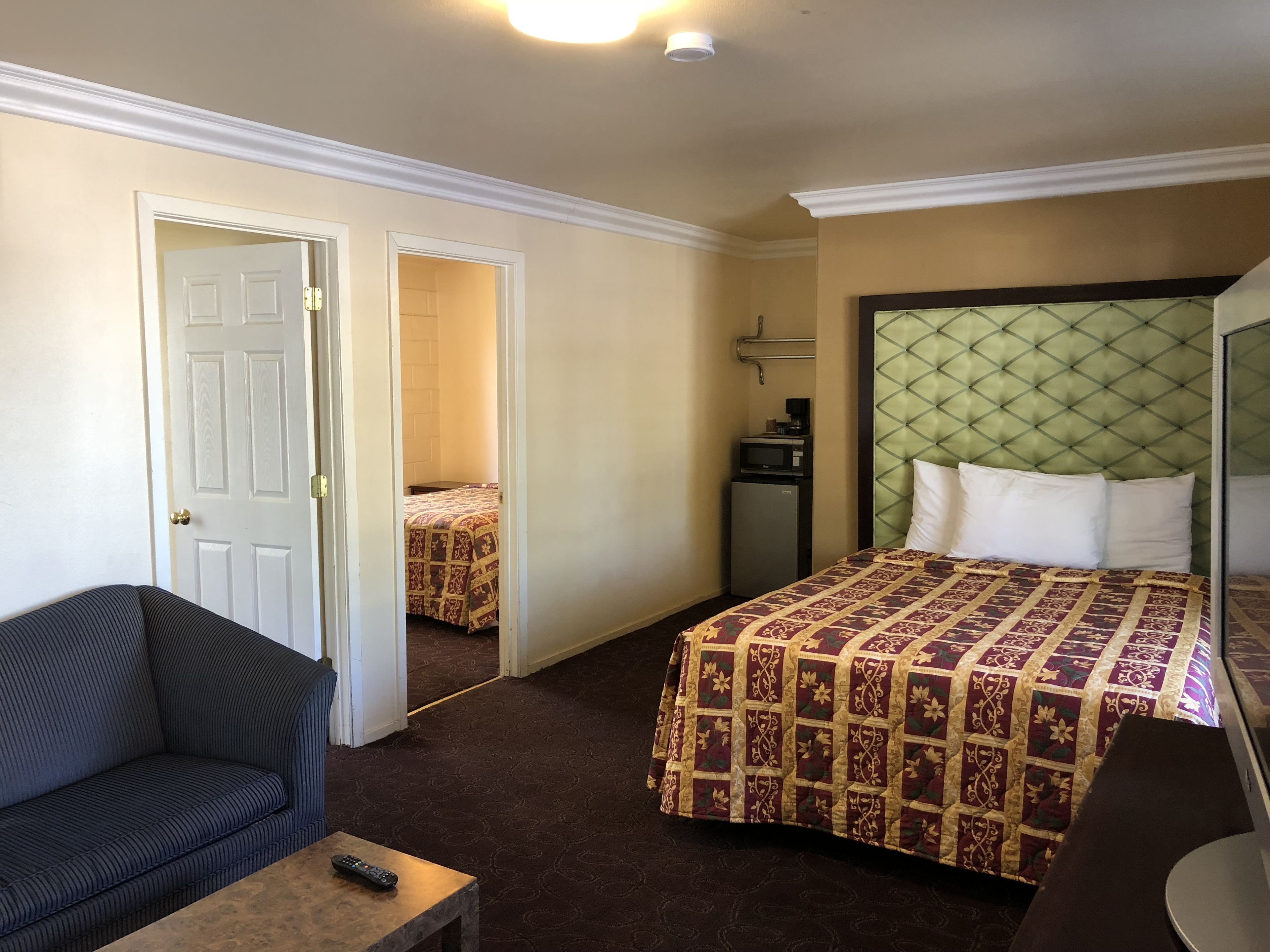 Habitación de hotel con cama, una mesita con un control remoto, una silla, un escritorio, un microondas y una puerta que da a otra habitación.