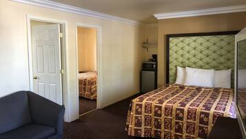 A hotel room with a bed, a small table with a remote control, a chair, a desk, a microwave, and a door leading to another room.