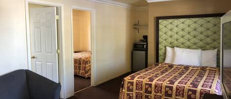 Habitación de hotel con cama, una mesita con un control remoto, una silla, un escritorio, un microondas y una puerta que da a otra habitación.