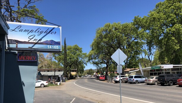 Ein Hotel namens 'Lamplighter Inn' mit einem 'VACANCY'-Schild, eine Straße mit geparkten Autos und ein Geschäft namens 'TURF & COLLECTIVE'.