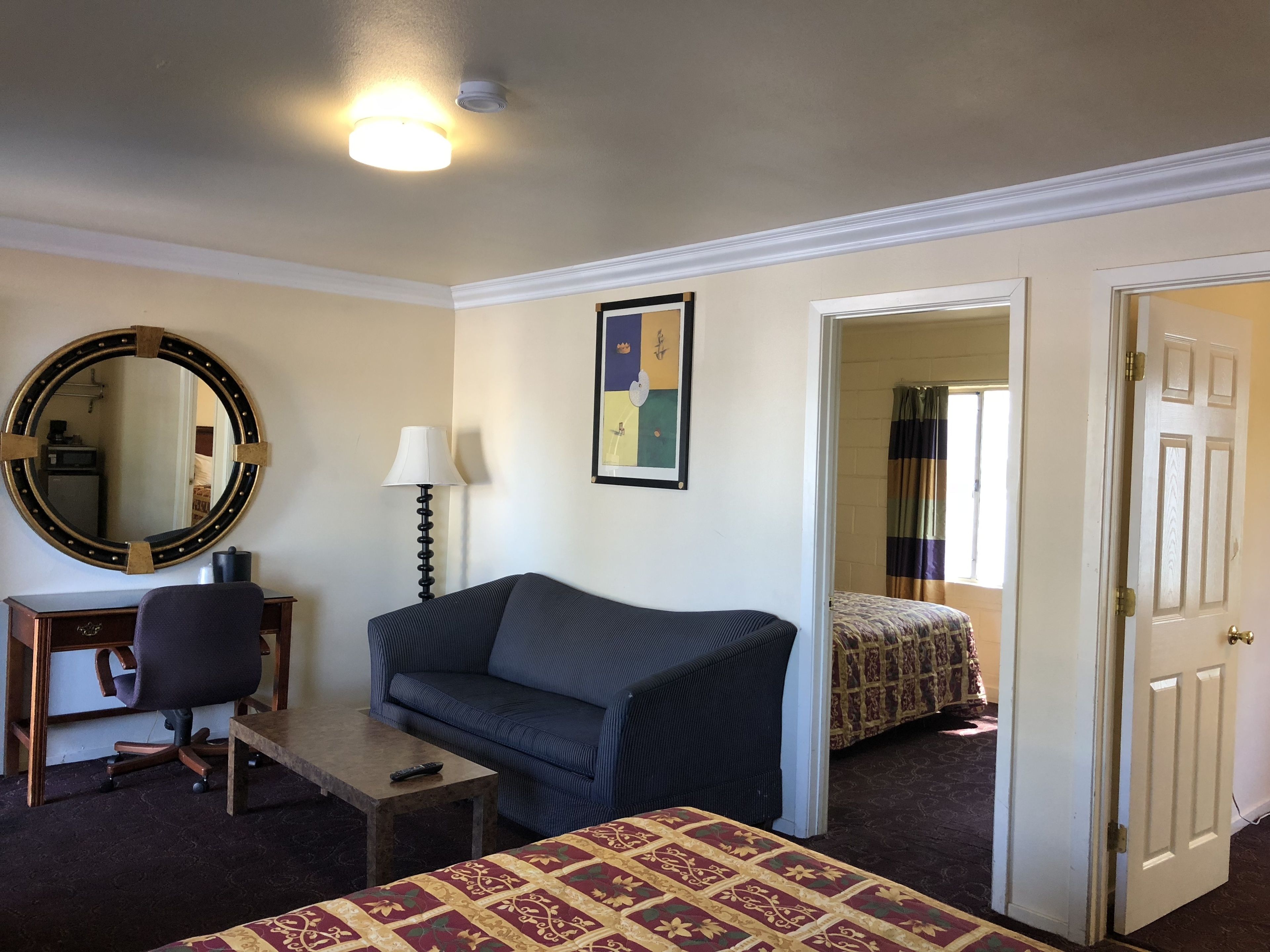 A hotel room with a desk, a couch, a bed, a mirror, a lamp, and a door leading to another room.